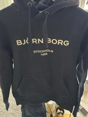 Svart hoodie från Björn Borg - Snygg svart hoodie från Björn Borg med stor vit logga och texten 'Stockholm 1956' på bröstet. Klassisk känguruficka framtill och justerbar huva med snören. Perfekt för en chill och sportig look. Mjuk bomullsblandning som känns skön mot huden.