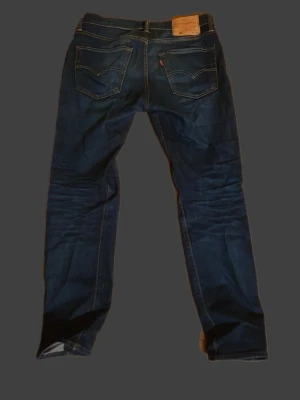 Levi's jeans - Klassiska Levi's jeans i mörkblå tvätt med raka ben och femficksmodell. Jeansen har tydliga kontrastsömmar, knappgylf och den ikoniska läderpatchen bak i midjan. Tillverkade i slitstark bomullsdenim som ger en tidlös look.