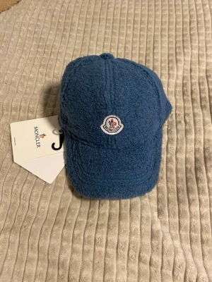Blå teddykeps från Moncler - Säljer en blå keps från Moncler i mjukt teddy-material. Kepsen har böjd skärm och ett broderat Moncler-märke framtill. Perfekt för dig som vill ha en unik och trendig accessoar som sticker ut. Justerbar baktill för bästa passform.
