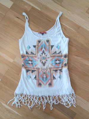 Vit bohemisk linne med fransar - Vit linne med tunna axelband och färgglatt aztekinspirerat mönster i blått, orange och svart. Toppen har fransar längst ner för en boho-vibe. 