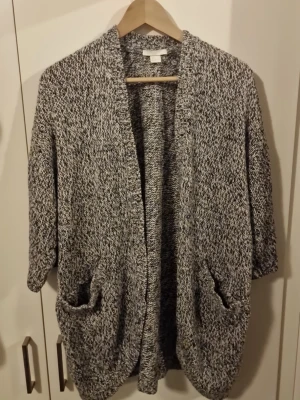 Gråmelerad stickad kofta med fickor - Mysig och oversized gråmelerad stickad kofta med två stora fickor framtill och avslappnad passform. Koftan har trekvartsärmar och öppen front utan knappar. Perfekt att slänga på sig över en t-shirt eller topp för en chill look.