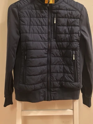 Mörkblå pufferjacka från Parajumpers juniorstorlek - Snygg mörkblå pufferjacka från Parajumpers juniorstorlek med quiltad framsida och dragkedjor på fickorna. Jackan har ribbade muddar och lång ärm, perfekt för kyligare dagar. Materialet är en mix av fleece och syntet för extra komfort.