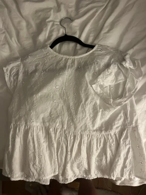 Vit blus med broderade blommor - Söt vit blus med broderade blommor och volang nedtill. Blusen har korta ärmar och v-ringning fram. Perfekt för varma dagar och ger en romantisk vibe till din outfit.