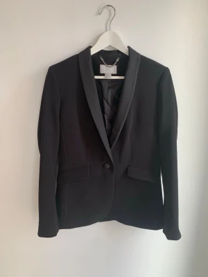 HM Kavaj 40 - Svart kavaj med en knapp framtill och detaljer i satin. Två fickor varav en fortfarande igensydd. Samfraktar.