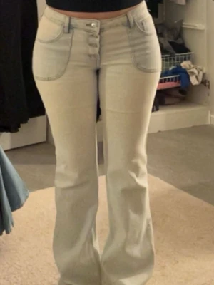Ljusblå lågmidjade flare jeans med knappar - Säljer ett par ljusblå jeans med raka ben och synliga knappar framtill. Jeansen har klassiska fem fickor och en cool, tvättad look. Perfekta för dig som gillar en avslappnad och trendig stil.