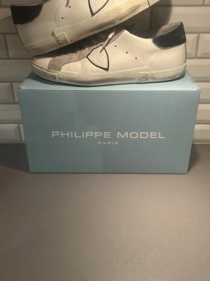 Vita Philippe Model sneakers med mockadetaljer - Snygga vita sneakers från Philippe Model med grå mockadetaljer och blåa partier vid hälen.  Skorna är i stl 44 och true to size. Köpta för 3200 kr. Knappt använda och inga defekter. Bara att höra av sig vid frågor eller funderingar😃Pris går att diskutera 