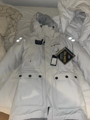 Vit sail racing jacka helt oanvänd som stått i garderoben i några år   - Snygg vit sail racing jacka, perfekt för kalla dagar. Jackan har huva, stora fickor med knappar, reflexdetaljer på axlarna och dragkedja framtill. Märkeslogga på bröstet och justerbar huva för extra skydd.