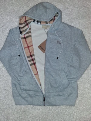 Grå hoodie från Burberry med dragkedja - Snygg grå hoodie från Burberry med klassiskt rutigt foder på insidan och dragkedja framtill. Hoodien har huva med dragsko, broderad logga på bröstet och två fickor. Tillverkad i mjukt bomullsmaterial för en skön känsla.