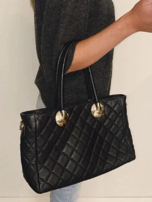 Svart quiltad handväska med gulddetaljer - Stilren svart Vera Pelle handväska i quiltad design med två rejäla handtag och snygga guldfärgade detaljer. Väskan har en klassisk rektangulär form och är tillverkad i skinnimitation. Perfekt för dig som vill ha en elegant och trendig accessoar till din outfit. Perfekt storlek för att få plats med allt du behöver och ger en stilren vibe till din outfit. 
