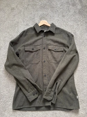 Mörkgrön overshirt i ullmix - Stilren mörkgrön overshirt med klassisk krage och två bröstfickor med knappar. Tillverkad i mjuk ullmix som ger en skön känsla och passar perfekt till lager på lager. Långa ärmar med knapp vid manschett och raka linjer för en clean look.                Bara att höra av sig vid eventuella frågor!