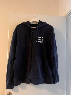 Mörkblå zip hoodie Peak Performance - Mörkblå hoodie med dragkedja från Peak Performance. Klassisk modell med huva och justerbara snören, samt fickor framtill. Diskret logga i vitt på bröstet. Perfekt för en avslappnad och sportig stil.