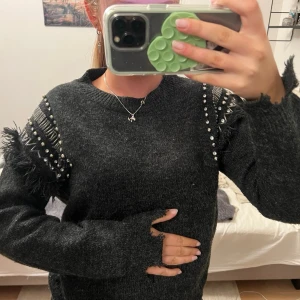 Mörkgrå stickad tröja med fransar och  nitar  - Säljer en mörkgrå stickad tröja med fransar och silverfärgade nitar på axlarna. Tröjan har en avslappnad passform, råa kanter vid ärmslut och nederkant samt rund halsringning. Perfekt för dig som gillar edgy detaljer och streetstyle.