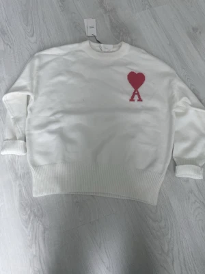 Vit stickad tröja från Ami Paris helt ny - Säljer en vit stickad tröja från Ami Paris med röd hjärtlogga och bokstaven A på bröstet. Tröjan har rund halsringning, ribbade muddar och nederkant. Perfekt för dig som gillar stilrena plagg med en twist.
