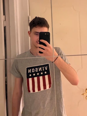 Två t-shirts från Vinson med tryck - Snygga gråa t-shirts från Vinson med stort tryck framtill i rött, vitt och blått. Trycket har texten VINSON och fem stjärnor ovanför röda och vita vertikala ränder. Klassisk passform och rund hals. Perfekt för en avslappnad stil. Båda tröjorna ingår i priset! Hör gärna av dig vid frågor eller liknande🙏