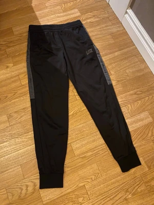 Svarta EA7 mjukisbyxor med logga - Snygga svarta mjukisbyxor från EA7 Emporio Armani med logga på benet och dekorativa band längs sidorna där det står 'EA7 Emporio Armani'. Byxorna har resår i midjan och muddar vid bensluten. Perfekta för en chill och sportig look. Tror att den är i storlek xs eller s 