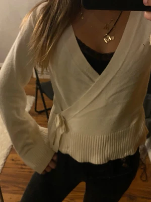 Krämvit omlottkofta med knytband - Säljer en krämvit stickad omlottkofta med knytband i sidan. Koftan har långa ärmar, ribbstickade muddar och en mjuk, mysig känsla. Perfekt att slänga över en topp för en chill look.