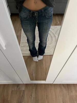 Blå lågmidjade bootcut jeans - Säljer ett par blå lågmidjade bootcut jeans med snygg tvätt och dubbla knappar framtill. Jeansen har klassisk femficksdesign och är tillverkade i slitstarkt denimtyg. Perfekta för en avslappnad och trendig look. 