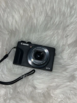 Canon G7X mark iii kompakt digitalkamera - Snygg och smidig svart Canon G7X digitalkamera med inbyggd Canon Zoom Lens 8.8-36.8mm f/1.8-2.8. Kameran har en klassisk rektangulär form och medföljande handledsrem. Perfekt för dig som vill ta grymma bilder och vlogga.