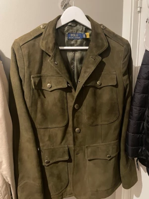 Olivgrön mockakavaj Polo Ralph Lauren - Snygg olivgrön trenchcoat i mockaliknande material från Polo Ralph Lauren. Jackan har fyra stora fickor framtill med lock och knappar, axelklaffar och klassisk krage. Perfekt för dig som gillar en stilren och tidlös look. Tyvär en liten spricka i insidan, inget som märks eller syns när man änvender den. Ny pris runt 10-12 tusen kr. 