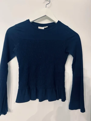 Mörkblå långärmad topp Gina Tricot - Mörkblå långärmad topp från Gina Tricot med smockad front och volangkant nedtill. Tillverkad i mjuk bomull, perfekt för en chill och stilren look. Rund halsringning och enkel design som är lätt att matcha.