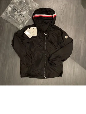 Svart vindjacka från Moncler - Snygg svart vindjacka från Moncler med huva och dragkedja framtill. Jackan har en röd och vit rand på huvan och Moncler-logga på ärmen. Tillverkad i lätt polyester, perfekt för blåsiga dagar. Klassisk passform och långärmad design.
