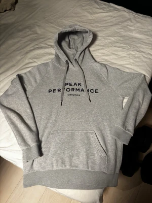 Grå hoodie från Peak Performance - Säljer en grå hoodie från Peak Performance med svart logga på bröstet. Klassisk modell med huva och dragsnören, samt en stor magficka. Tillverkad i mjukt bomullsmaterial och har ribbade muddar vid ärmar och nederkant. Perfekt för en avslappnad och sportig stil. Storlek junior L så ungefär S/M i vanlig storlek 
