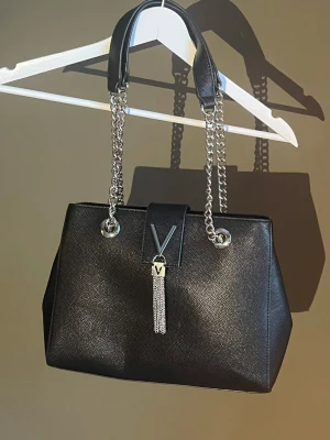 Valentino väska med kedjehandtag - Snygg svart väska från Valentino med silverfärgade kedjehandtag och dekorativ V-detalj framtill. Väskan har en elegant tofs i metall och är tillverkad i skinnliknande material. Perfekt accessoar för att lyfta din outfit. Köpt från Valentino, använts ett par gånger 🖤