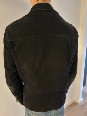 Svart mocka bikerjacka med dragkedjor - Snygg svart bikerjacka i mocka med flera dragkedjor framtill och klassisk krage med tryckknappar. Jackan har lång ärm och en cool, lite tuff look som passar perfekt till jeans. Perfekt för dig som vill ha en stilren och trendig skinnjacka.