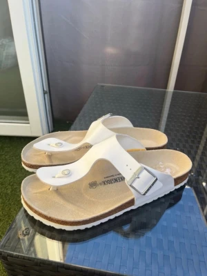 Vita Birkenstock sandaler med spänne - Säljer ett par klassiska vita Birkenstock sandaler med tårem och justerbart spänne i metall. Sandalerna har en bekväm fotbädd och platt sula, perfekta för sommaren. Stilsäker design som funkar till det mesta.