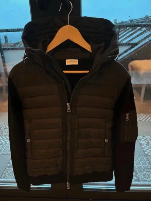 Svart cardigan jacka från Moncler - Svart cardigan jacka från Moncler med quiltade partier och stickade ärmar. Jackan har huva, tvåvägsdragkedja och fickor med dragkedja. Perfekt för dig som vill ha en stilren och sportig look.
