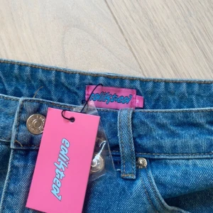 Blå jeanskjol från Edikted - Trendig blå jeanskjol från Edikted med klassiska fickor bak och slitna detaljer. Kjolen har en kort passform och stängs med knapp och dragkedja framtill. Perfekt för dig som gillar en avslappnad och cool stil.