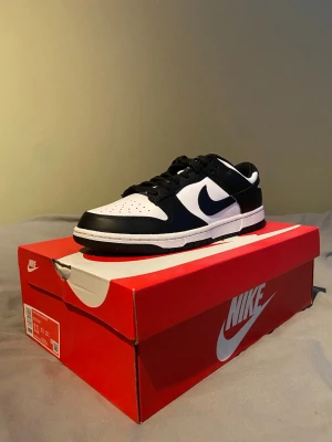 Nike Dunk Low svart/vit sneakers - Nike Dunk Low sneakers i svart och vitt med klassisk låg siluett. Skon har svart snörning, vit tåbox och sidor, samt svarta overlays och Swoosh. Tillverkad i skinn med platt sula och rund tå. Perfekt för dig som gillar stilrena och tidlösa sneakers.