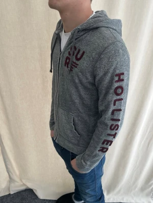 Grå zip hoodie från Hollister - Snygg grå zip hoodie från Hollister med stor logga i vinrött längs ena ärmen och tryck på bröstet. Strl M. Personen på bilden är 184 cm lång. Skicka prisförslag. Kolla min profil för liknande kläder.
