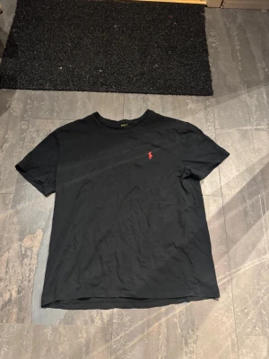 Polo Ralph lauren  - Klassisk svart t-shirt från Polo Ralph Lauren med den ikoniska röda logobrodyrn på bröstet. T-shirten har rund hals och korta ärmar, tillverkad i mjuk bomull för en skön känsla. Perfekt basplagg med stilren design.