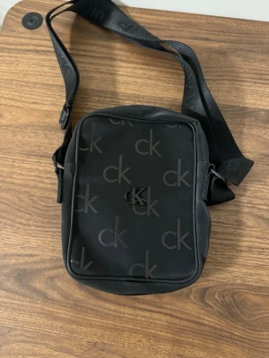 Svart axelväska från Calvin Klein - Snygg svart axelväska från Calvin Klein med justerbar rem och diskret ck-logga över hela väskan. Väskan har dragkedja både fram och upptill, perfekt för att hålla koll på dina grejer. Stilren och enkel design som passar till det mesta.