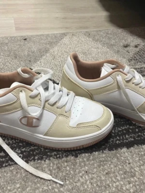 Vita och beige sneakers från Champion - Snygga sneakers från Champion i vitt och beige med ljusbruna detaljer. Skorna har klassisk snörning, rund tå och platt sula. Perfekta för dig som gillar en clean och stilren look. Champion-logga på sidan och under sulan.