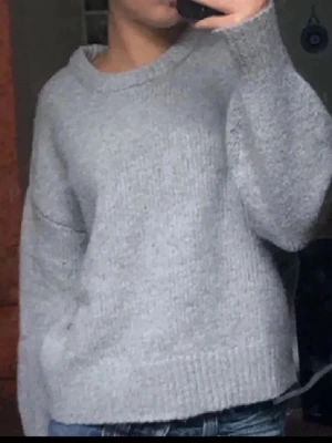 Grå stickad tröja med rund hals - Mysig grå stickad tröja med rund halsringning och ribbade muddar vid ärmslut och nederkant. Obs tröjan har några få små rosa fläckar vid halsringen skriv för bilder