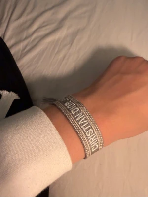 Dior armband - Snyggt grått armband från Christian Dior med broderad logga i vitt. Armbandet är gjort i textil och har justerbar knytning med tofsar i ändarna. Perfekt accessoar för att lyfta din outfit med en lyxig touch.