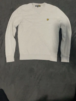 Ljusgrå sweatshirt från Lyle & Scott - Ljusgrå sweatshirt från Lyle & Scott med klassisk rund halsringning och gul logobrodyr på bröstet. Tröjan har ribbade muddar vid ärmslut och nederkant, perfekt för en clean och enkel stil.