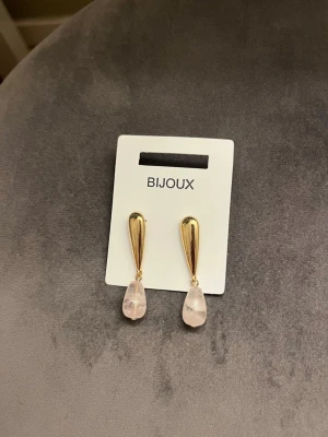 Guldörhängen med rosa droppar Bijoux - Snygga örhängen från Bijoux med droppformad design i guldmetall och ljusrosa pärlliknande hängen. Perfekta för att ge din look en elegant touch. Kombinerar modern stil med klassiska detaljer.