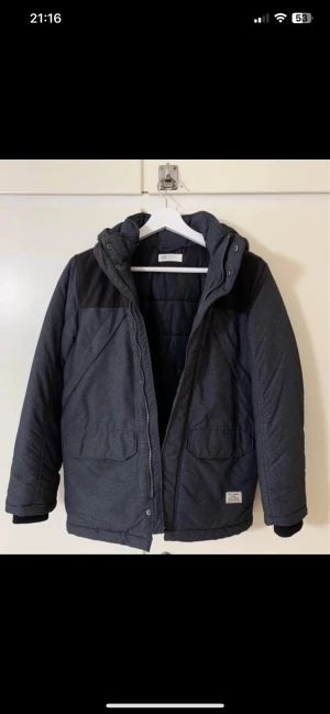 Mörkgrå dunjacka från H&M - Snygg mörkgrå dunjacka från H&M med svarta detaljer över axlarna och huva. Jackan har två stora fickor framtill, dragkedja och tryckknappar. Perfekt för kalla vinterdagar och har en stilren look.