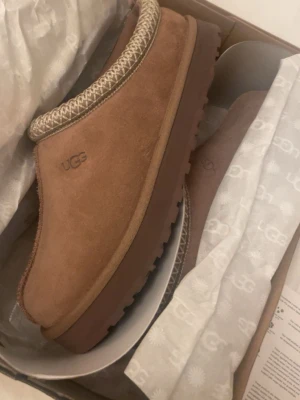 Bruna UGG tofflor i mocka - Säljer ett par snygga bruna UGG tofflor i mjuk mocka med grov sula och dekorativt band runt öppningen. Perfekta för att hålla fötterna varma och sköna hemma eller ute. Klassisk UGG-logga på sidan och riktigt trendig look.