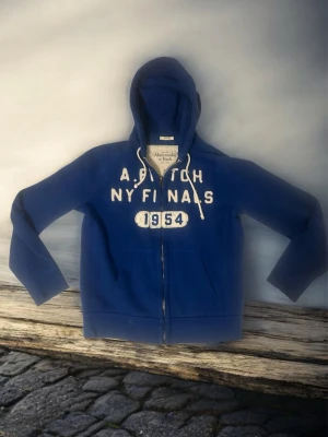 Blå hoodie Abercrombie & Fitch - Blå zip-hoodie från Abercrombie & Fitch med vit text 'A&Fitch NY Finals 1954' på bröstet. Utrustad med huva och vita dragsnören samt fickor framtill. Mjuk bomullskänsla och ribbade muddar vid ärmslut och nederkant.