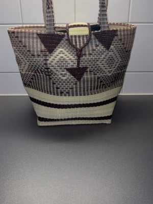 Brun och beige flätad shoppingväska - Snygg shoppingväska i flätad plast med geometriskt mönster i brunt och beige. Väskan har två handtag och en smart knäppning upptill. Perfekt för att bära med sig allt man behöver under dagen.