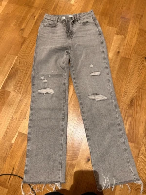 Gråa raka jeans från Zara - Säljer ett par gråa raka jeans från Zara med slitna detaljer och fransade benslut. Jeansen har hög midja, klassisk femficksdesign och är tillverkade i denim. Perfekta för en avslappnad och trendig look.