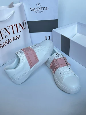 Valentino Garavani vita sneakers - Snygga vita sneakers från Valentino Garavani med ljusrosa band över vristen dekorerat med ikoniska nitar. Skorna har en låg siluett, vita snören och detaljer i läder. Perfekta för dig som vill ha en exklusiv och trendig look.