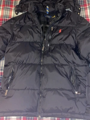 Svart dunjacka från Polo Ralph Lauren - Svart dunjacka från Polo Ralph Lauren med röd broderad logga på bröstet. Jackan har dragkedja framtill, två sidofickor med dragkedja och hög krage. Klassisk puffer-modell som håller dig varm under kalla dagar. Ett litet hål i en av fickorna