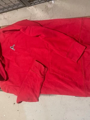 Röd Jordan hoodie med broderad logga - Snygg röd hoodie från Jordan med den klassiska broderade loggan på bröstet. Hoodien har en stor ficka framtill och ribbade muddar vid ärmsluten. Perfekt för dig som gillar streetwear och sportig stil. Materialet känns mjukt och bekvämt.