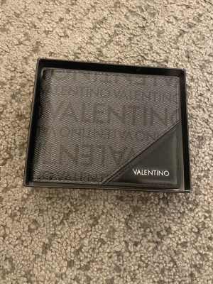 Svart plånbok från Valentino - Snygg svart plånbok från Valentino med diskret logomönster över hela ytan och en stilren detalj i hörnet med VALENTINO-tryck. Plånboken är tillverkad i läder och har en klassisk, fyrkantig form. Perfekt för dig som gillar stilrena accessoarer. Du får även med boxen den är helt ny plånboken inte använd en gång 