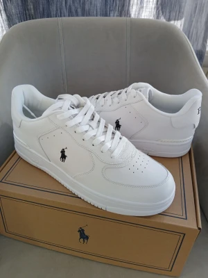 Nya Polo Ralph Lauren sneakers strl 45 - Stilrena vita sneakers från Polo Ralph Lauren med klassisk låg siluett. Skorna har en vit ovandel i läder med den ikoniska Polo-loggan broderad på sidan och diskret branding på hälen. Perfekta för en clean och sportig look. Nypris 1845 kr